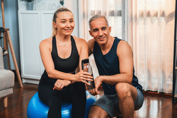 Fototapeta na wymiar Athletic and sporty senior couple portrait in sportswear sitting on sofa with fitness water bottle as home exercise concept. Healthy fit body lifestyle after retirement. Clout