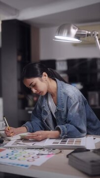 Asian female architect meticulously designs products on her computer, selecting color tones, contributing to a modern office setting. Her creations bring vibrancy
