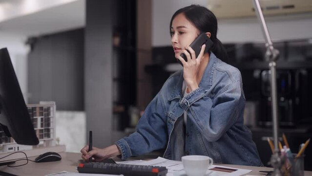 Asian female architect, focused and attentive, engages in a phone conversation while working on computer-aided design for products in a modern office setting.
