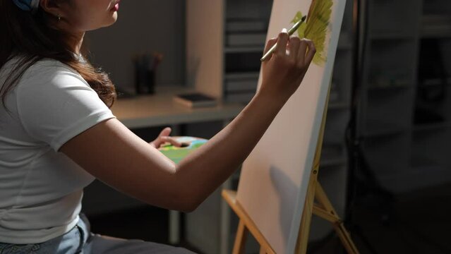 A young Asian female artist and painter paints with a paintbrush with painting on an easel in the art studio with acrylic paints on canvas. Lifestyle hobby ideas.