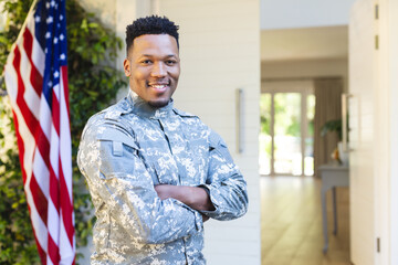 African american male soldier standing with arms crossed outside home with usa flag, copy space
