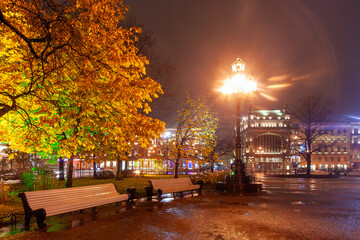 Obraz premium Evening view of empty benches in the city park and beautiful buildings in the historical center of St. Petersburg. Rainy autumn weather. Nevsky Prospekt, St. Petersburg, Russia. Night city.