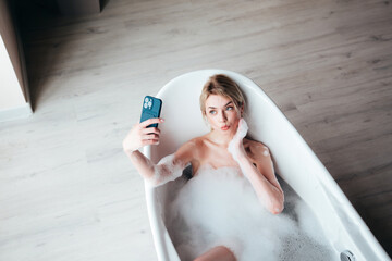 Young beautiful sexy woman having fun while lying in bathtub full of foam at home. Charming smiling...