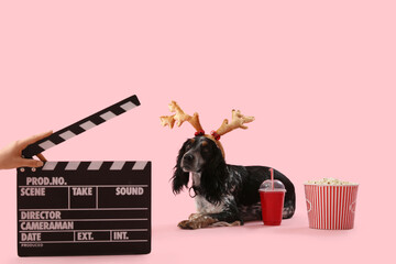 Cute cocker spaniel in reindeer horns with soda, popcorn and movie clapper on pink background