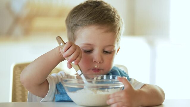 Baby eats cereal porridge. Bboy is holding spoon in his hand. Child sits at table and eats porridge.Family breakfast in kitchen. Feeding child at table.Child proper nutrition.Breakfast in kindergarten