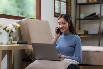Attractive Asian woman sitting talking on phone and laptop computer in living room at home.