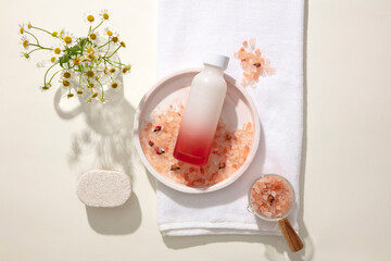 An unlabeled cosmetic bottle is displayed on ceramic plates with pink salt. Daisies, a towel and props are decorated around. Mockup for product advertising. New product launch concept.