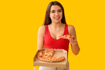 Young woman eating piece of tasty pizza and holding cardboard box on yellow background