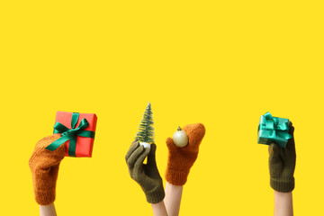 Female hands in warm gloves with Christmas gifts and decorations on yellow background