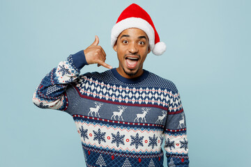 Young excited man wear knitted sweater Santa hat posing doing phone gesture like says call me back isolated on plain pastel blue background. Happy New Year 2024 celebration Christmas holiday concept.