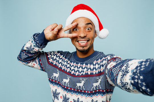 Close up young man wearing sweater Santa hat posing doing selfie shot pov on mobile cell phone show v-sign isolated on plain blue background. Happy New Year 2024 celebration Christmas holiday concept.