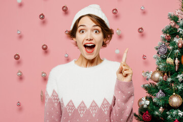 Merry proactive young woman wears white sweater hat posing holding index finger up with great new idea isolated on plain pastel pink background. Happy New Year celebration Christmas holiday concept.