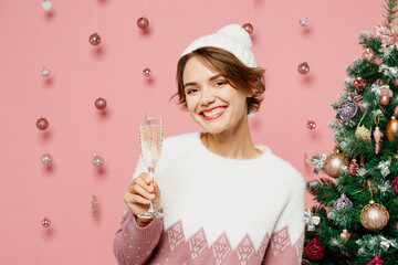Merry young woman wears white sweater hat posing hold in hand wineglass with champagne beverage isolated on plain pastel pink background studio. Happy New Year celebration Christmas holiday concept.