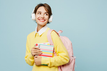 Obraz premium Young fun woman student wear casual clothes yellow sweater backpack bag hold books listen to music in headphones look aside isolated on plain blue background. High school university college concept.