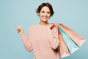 Young woman wear beige knitted sweater casual clothes hold in hand paper package bags after shopping point aside isolated on plain light pastel blue cyan background. Black Friday sale buy day concept. - obrazy, fototapety, plakaty
