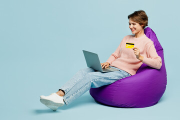 Full body young smiling woman she wearing beige knitted sweater casual clothes sit in bag chair using mobile cell phone hold credit bank card doing online shopping isolated on plain blue background.