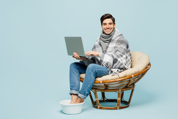 Full body young ill sick man wrapped in plaid sit in chair keeps feet in basin use mobile cell phone isolated on plain blue background. Healthy lifestyle virus treatment cold season recovery concept.
