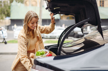 Young smiling happy customer woman wearing casual clothes putting paper bag into car trunk shopping...