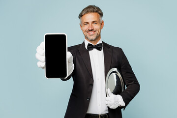 Adult barista male waiter butler man wear shirt black suit bow tie uniform hold tray use show blank screen mobile cell phone work at cafe isolated on plain blue background Restaurant employee concept