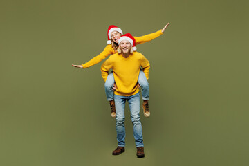 Full body merry young couple two friends man woman wear sweater Santa hat posing giving piggyback ride joyful, sit on back show ok isolated on plain green background. Happy New Year Christmas concept. - obrazy, fototapety, plakaty