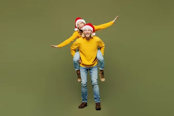 Fotobehang Full body merry young couple two friends man woman wear sweater Santa hat posing giving piggyback ride joyful, sit on back show ok isolated on plain green background. Happy New Year Christmas concept. © ViDi Studio