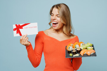 Young happy excited woman wear orange casual clothes hold store gift coupon voucher card eat raw fresh sushi roll served on black plate Japanese food isolated on plain blue background studio portrait.