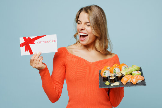 Young happy excited woman wear orange casual clothes hold store gift coupon voucher card eat raw fresh sushi roll served on black plate Japanese food isolated on plain blue background studio portrait. - Powered by Adobe