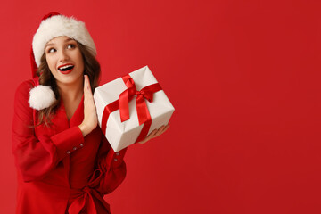 Surprised young woman in Santa hat with Christmas gift box on red background
