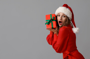 Surprised young woman in Santa hat with Christmas gift box on grey background