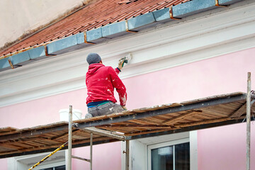 Worker on scaffolding paint facade wall of historic building. Utility worker paints building facade. Repair and restoration building facade. Man painting house wall with paintbrush - obrazy, fototapety, plakaty