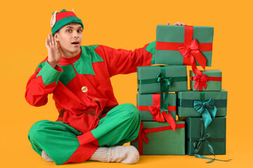 Handsome young man in elf's costume with gift boxes on yellow background