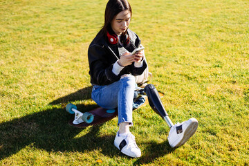 Fototapeta premium Young woman student with prosthetic leg sitting on green grass in university campus and using smartphone. Disabled woman with bionic leg in public park. Woman with prosthesis equipment using phone