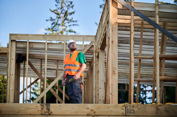 Carpenter building wooden frame two-story house. Bearded man in glasses holding a level, wearing protective helmet, overalls and orange vest. Concept of modern ecological construction.