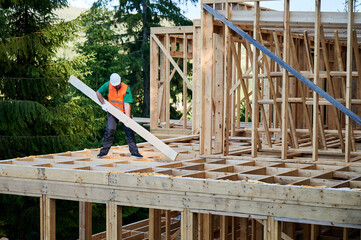 Carpenter builds wooden frame house near woods. Man holds large joist, wearing work clothes and...