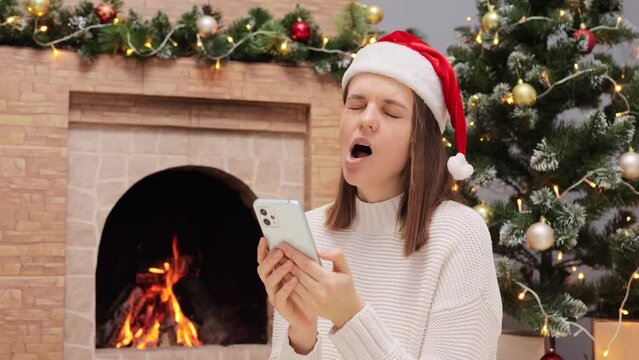 Sleepy woman wearing warm sweater and Santa Claus hat sitting in living room near fireplace and Christmas tree reading news in network yawning scrolling online long hours.