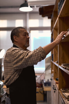 Bearded Man In Checked Shirt Choosing Selecting Instrument To Make Clothes From Leather Working Process. Side View Shot Tailor Putting Tools On Shelf. Man Tiding Up Cleaning Favorite Job