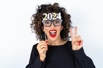 Young beautiful woman with curly hair wearing black dress over white studio background holding a Happy New year 2024 banner glasses and making victory sign. 