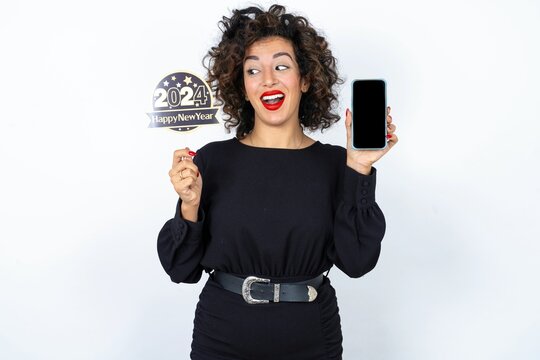 Young beautiful woman with curly hair wearing black dress, holding happy new year 2024 message showing blank screen picture. Woman looking aside at her phone.