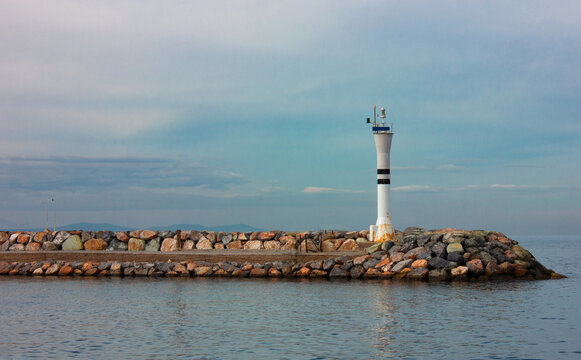 A lighthouse on a stone port or harbor under the blue and cloudy sky. Calm sea and beautiful sky seascape image.