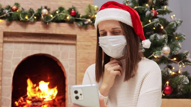 Adult woman with brown hair wearing Santa Claus hat and medical mask sitting in decorated living room with Christmas tree and burning fireplace talking via smartphone social distance virus disease.