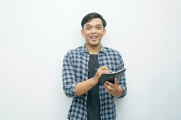 Portrait of young Asian Indonesian man smiling when writing on small notebook with ballpoint pen...
