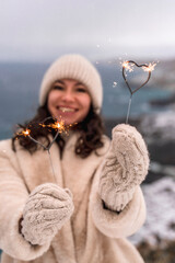 Outdoor winter portrait of happy smiling woman, light faux fur coat holding heart sparkler, posing...