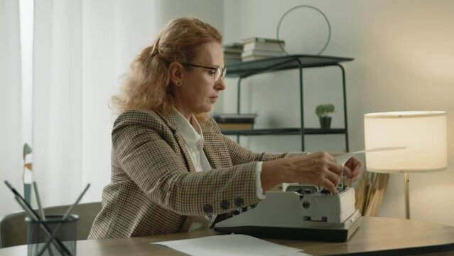 Experienced blonde typist putting sheet of paper into vintage mechanical typewriter, starting typing, sitting at table in light room with modern interior. High quality 4k footage