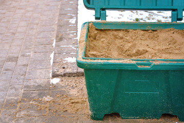 Naklejka premium Grit bin with sand for treatment paving slabs from snow and ice, close up. Sand box for improve traction on snowy, icy sidewalk, road maintenance in winter season. Selective focus