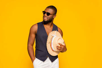 Smiling young african american fashionable man wears suit vest showing torso, sunglasses and hat at studio isolated over yellow background. Stylish male, fashion concept.