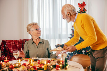 A senior man is serving his wife and bringing food at christmas table on christmas and new year at home.