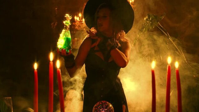 fantasy woman witch creates magic in dark gothic room full smoke candle fire. evil mystical girl holds in hands glowing bottle poison potion. Carnival costume witch magician black dress pointed hat 4k