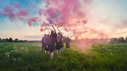 Graduates in costume walk with a smoky multi-colored smoke at sunset - obrazy, fototapety, plakaty