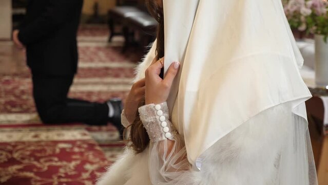 During a church wedding, the bride prays before the icon, standing on her knees with a scarf on her head. Church wedding traditions.