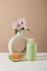 An unlabeled cosmetic bottle is placed next to a pomelo and a flower vase on a pastel background....